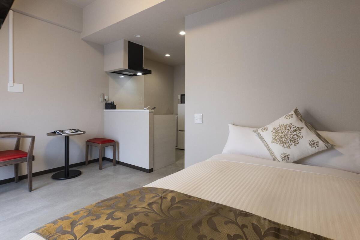 A hotel room with a bed, a small desk with a chair, a kitchenette, and a bathroom visible in the background.