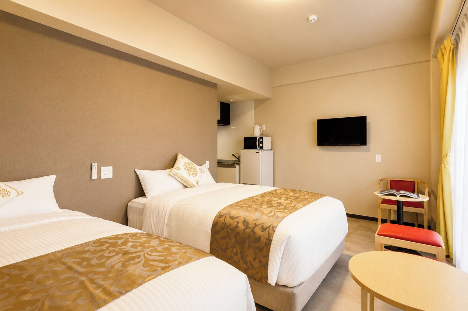 A hotel room with two beds, a small kitchenette, a TV, and a table with a chair.