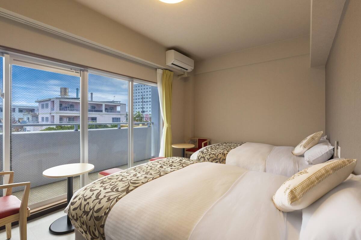 A hotel room with a large window, a bed with patterned bedding, a small round table, and a chair.
