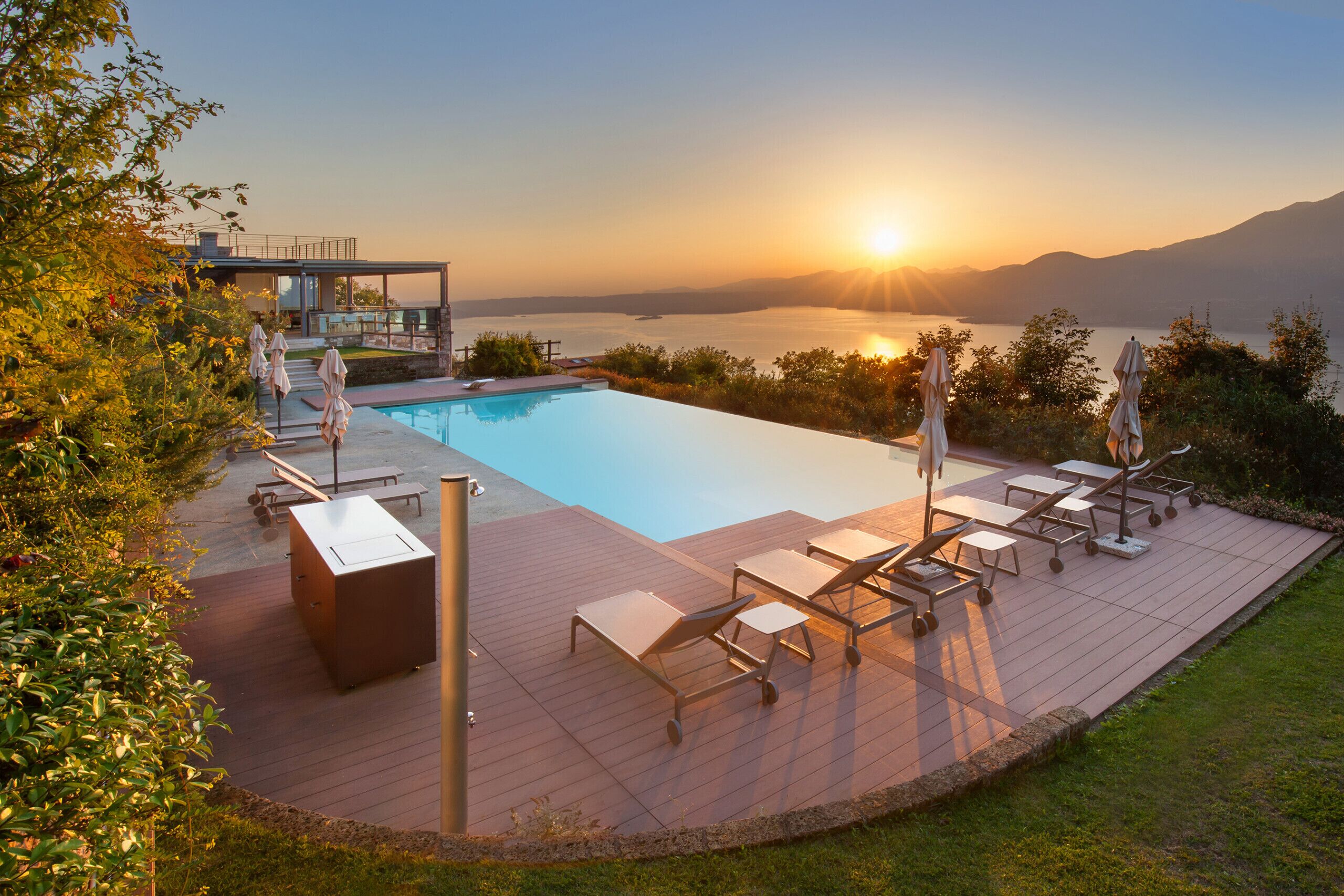 A pool area with lounge chairs and umbrellas, overlooking a body of water and mountains during sunset.