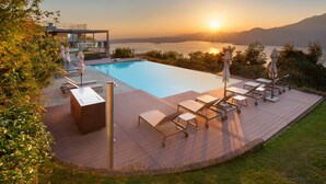 Zona de piscina con sillones de descanso y sombrillas, con vistas a un cuerpo de agua y montañas al atardecer.