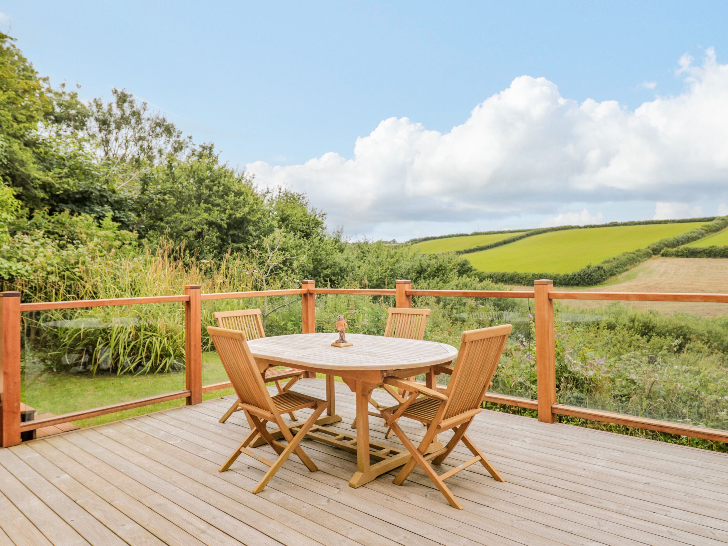 Una terraza de madera con una mesa redonda y cuatro sillas, con vistas a un paisaje verde con campos y árboles.