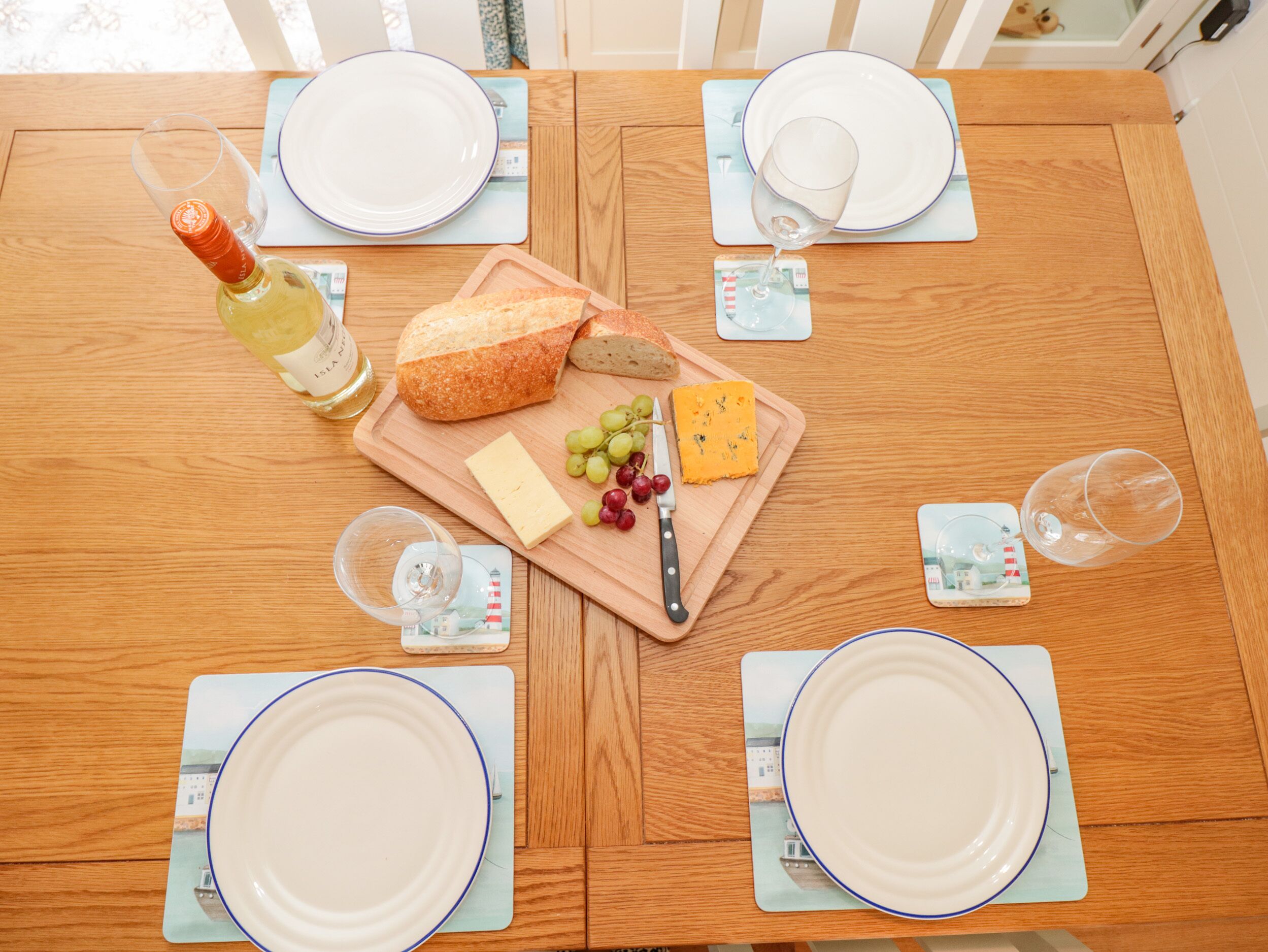 Une table de salle à manger en bois, mise pour quatre personnes, avec des assiettes, des verres, une bouteille de vin, une miche de pain, du fromage et des raisins.