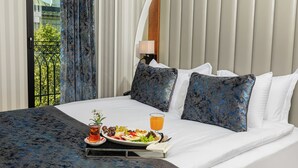 Cama bem arrumada com roupa de cama branca e almofadas estampadas em azul, uma bandeja com café da manhã incluindo frutas e um copo de suco de laranja, e vista para o verde através de uma janela com cortinas.