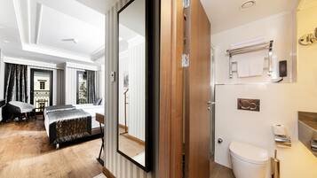 Ett hotellrum med en stor spegel, en säng med ett metalliskt överdrag, ett trägolv, ett badrum med en toalett och en handduksställning.
