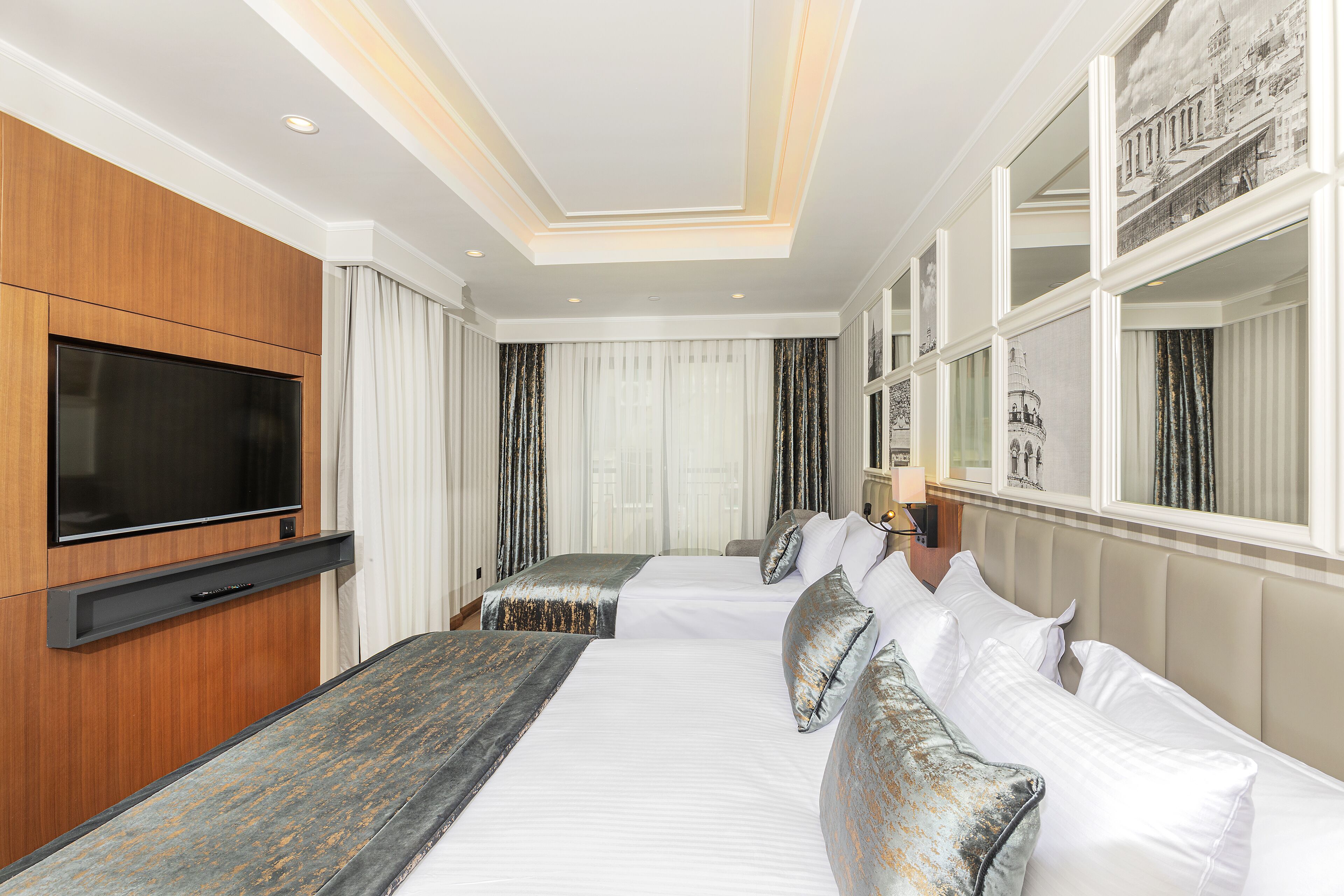 A hotel room with a large flat-screen TV mounted on the wall, a bed with white bedding and decorative pillows, a wooden headboard, and a large window with curtains.