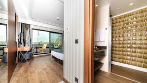 Quarto de hotel moderno com piso de madeira, cama com vista, área de estar com mesa e cadeiras e banheiro com parede de azulejos únicos.