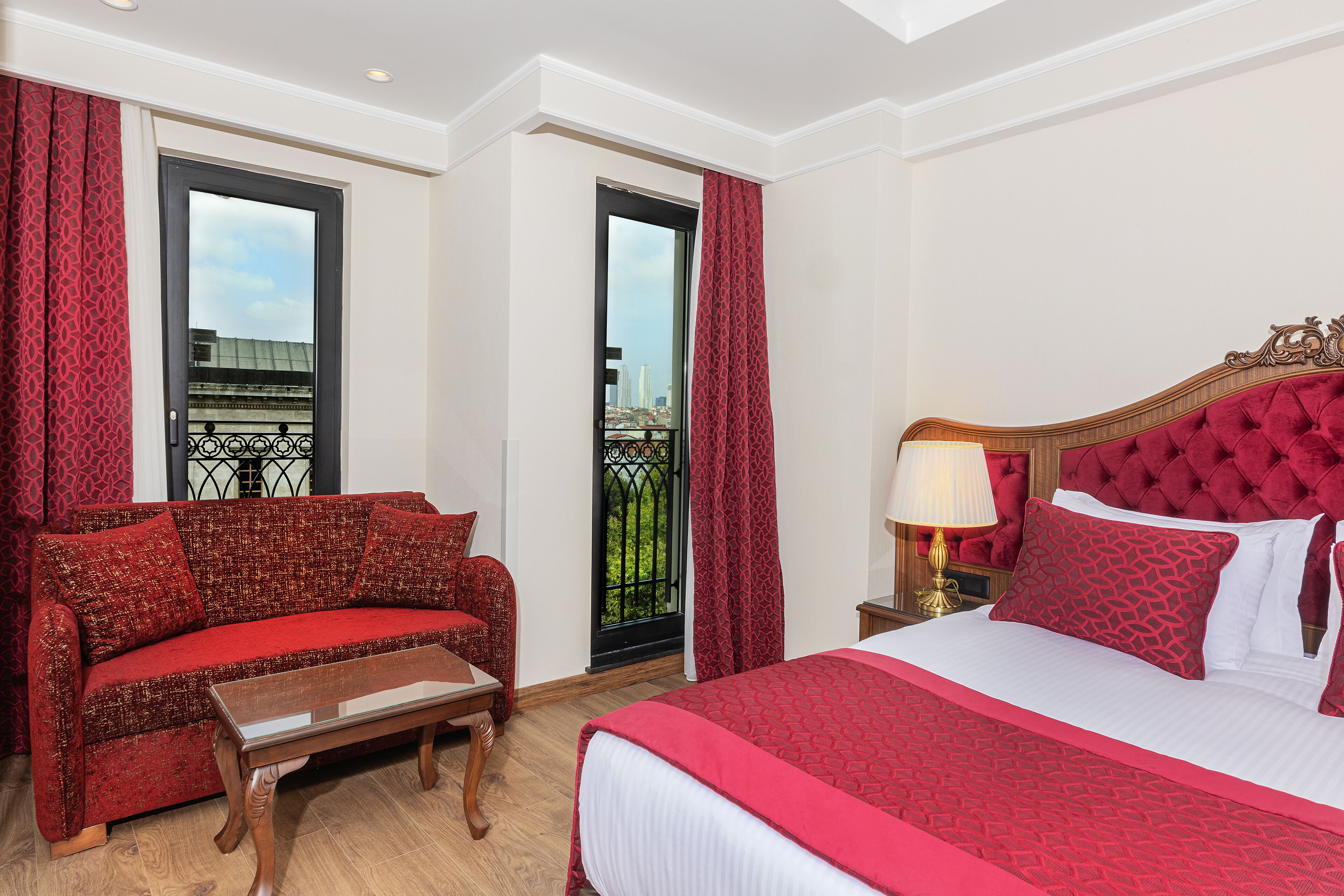 Une chambre d’hôtel avec un canapé rouge, une table basse en bois, un grand lit recouvert d’une couverture rouge et une table de chevet avec une lampe.
