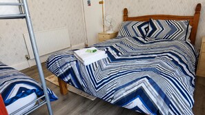 Un llit individual amb un estovat estriat, un panell frontal de fusta, una tauleta de nit amb una làmpada i un radiador a la paret.