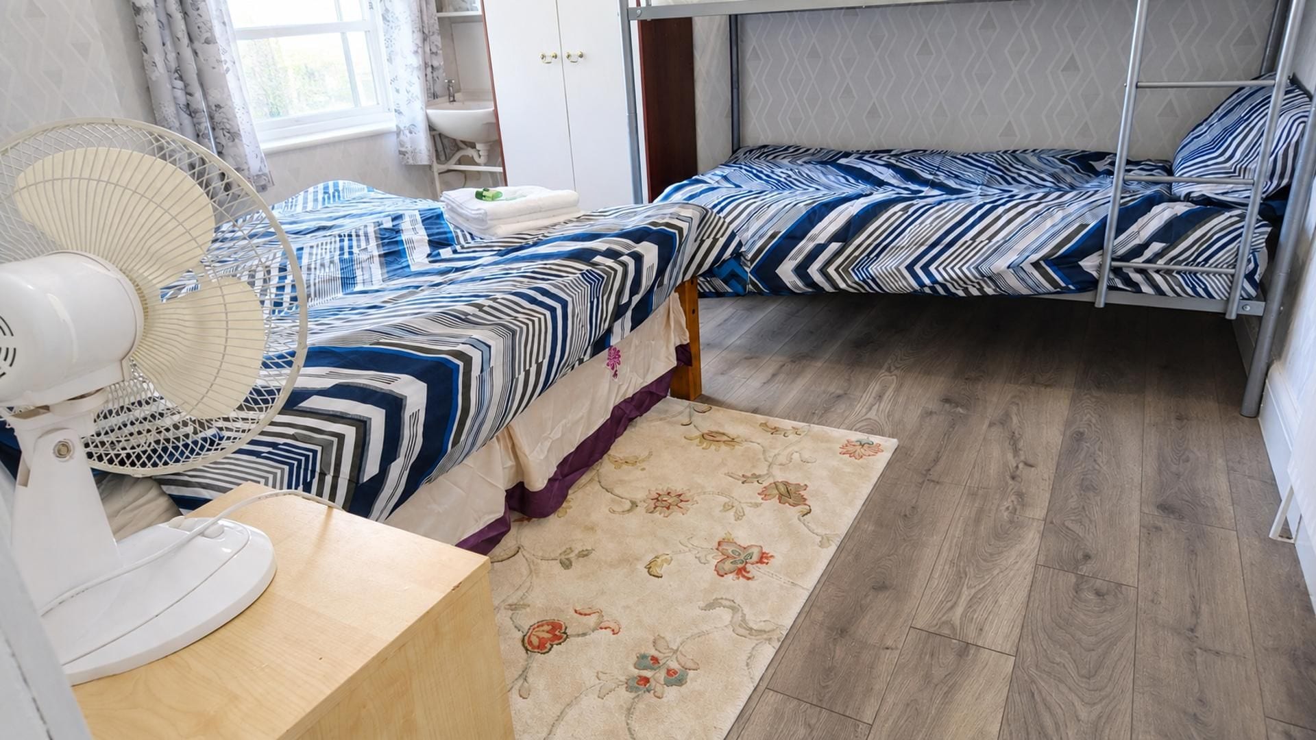 A room with two beds, a fan, a sink, and a window with curtains.