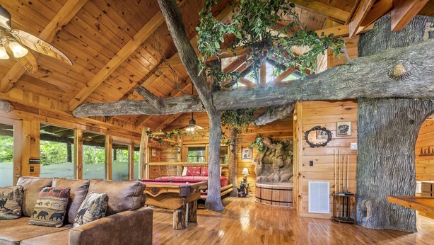 A log cabin interior with a wooden ceiling, a large tree trunk centerpiece, a sofa with animal-themed cushions, and a billiards table.