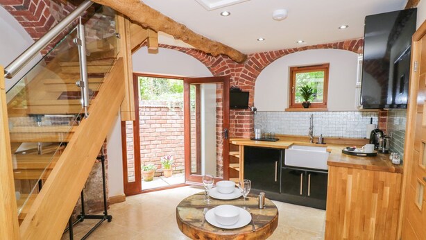 A modern kitchen with a wooden table set for two, a brick wall, and a staircase leading to another level. - The Hyde Dovecote Kinver, pet Friendly Holiday let (Stourbridge)