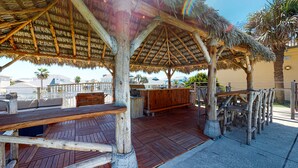 Eine Holzterrasse mit einem strohgedeckten Unterstand, Sitzgelegenheiten im Freien und einem Grillbereich.