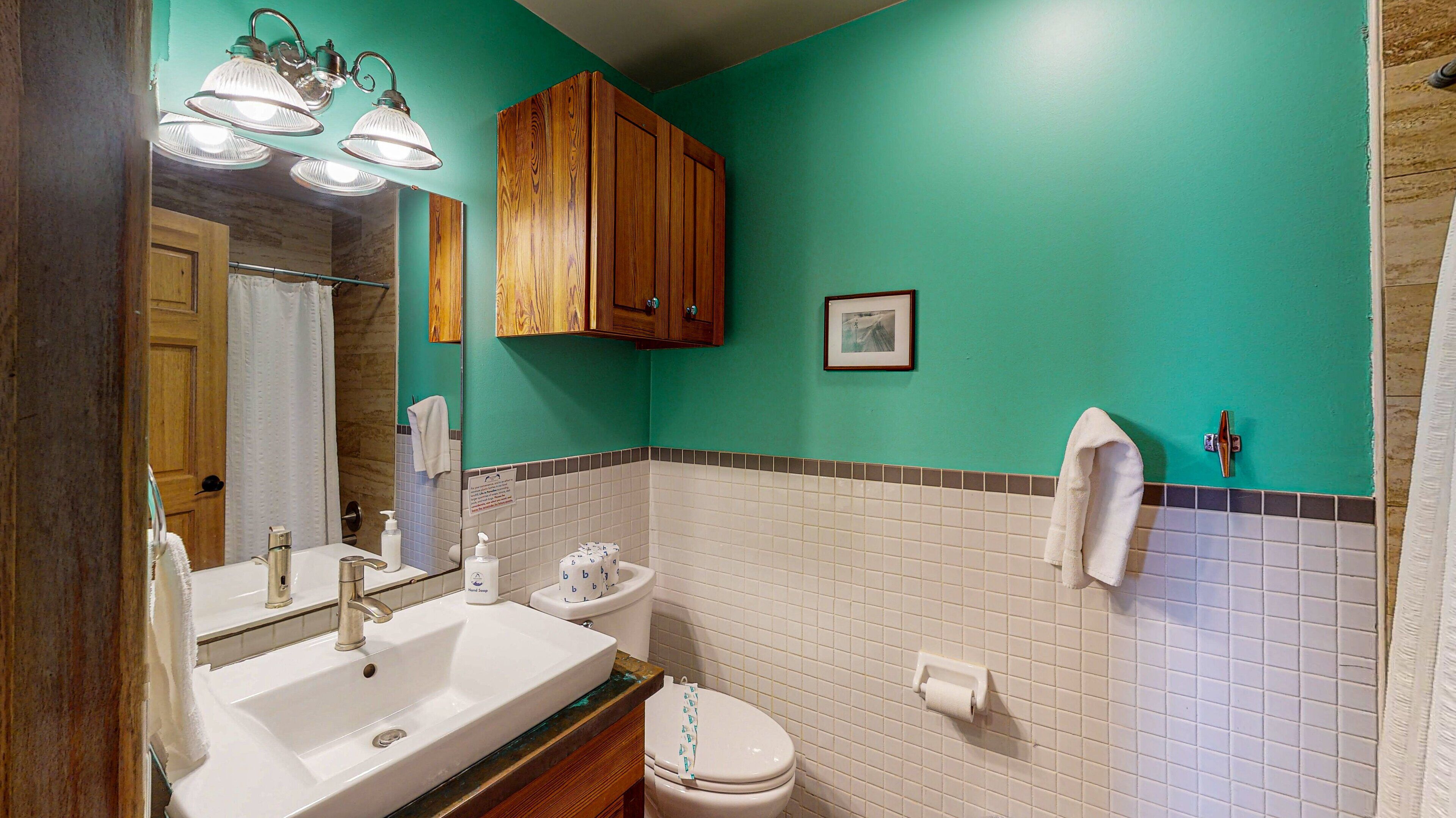 Ein Badezimmer mit Waschbecken, Toilette und Spiegel. Oberhalb des Spiegels befindet sich ein Holzkorpus und ein Handtuch hängt an der Wand.