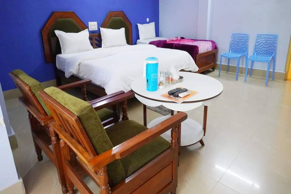 A hotel room with two beds, a round table with a blue water bottle, and two blue chairs.