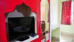 Una habitación con un armario rojo, un televisor sobre una estantería blanca, un espejo y una cortina roja con diseño.