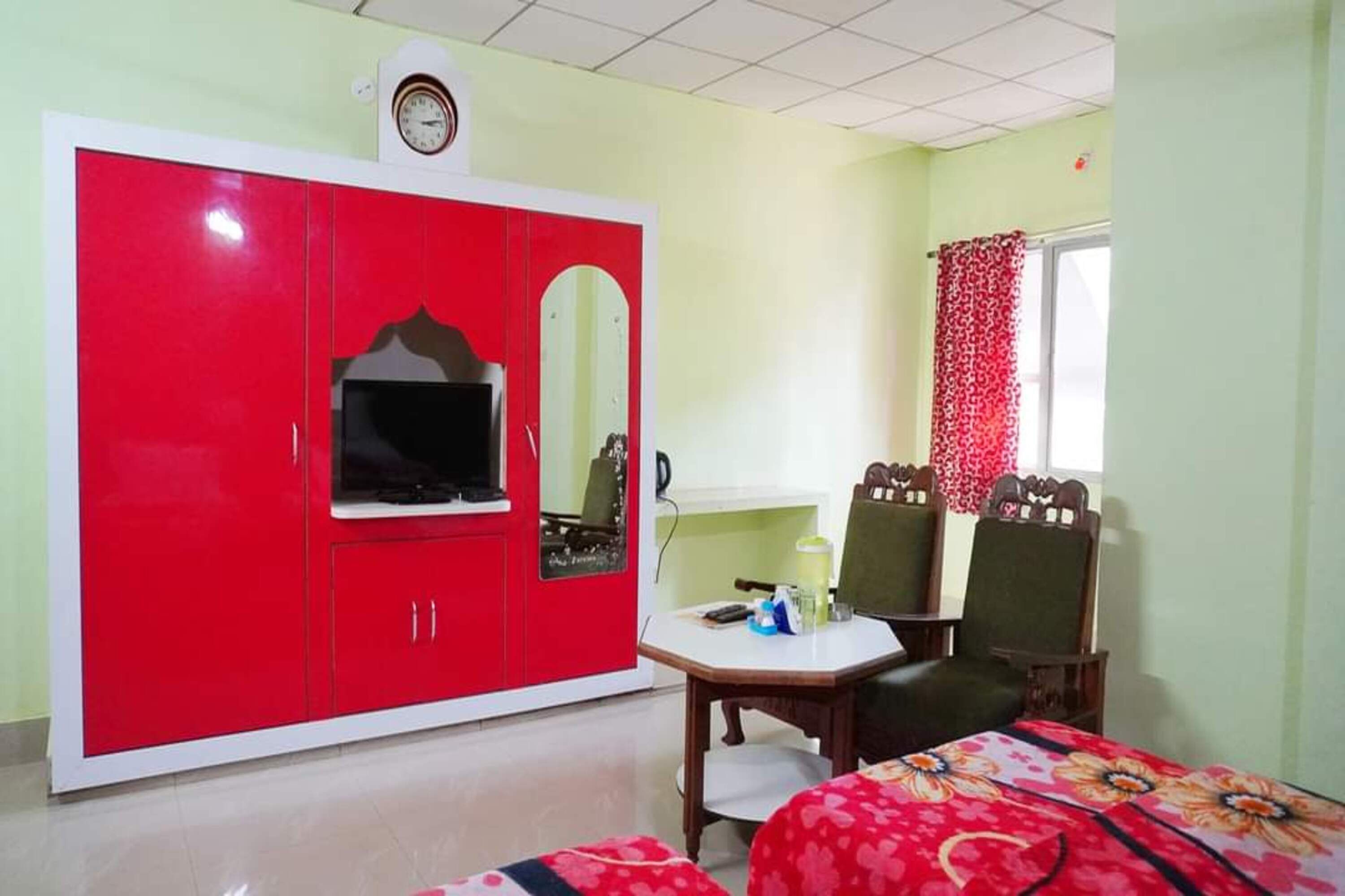 A room with a red wardrobe, a small table, two chairs, a bed with a floral bedspread, a clock on the wall, and a window with red curtains.