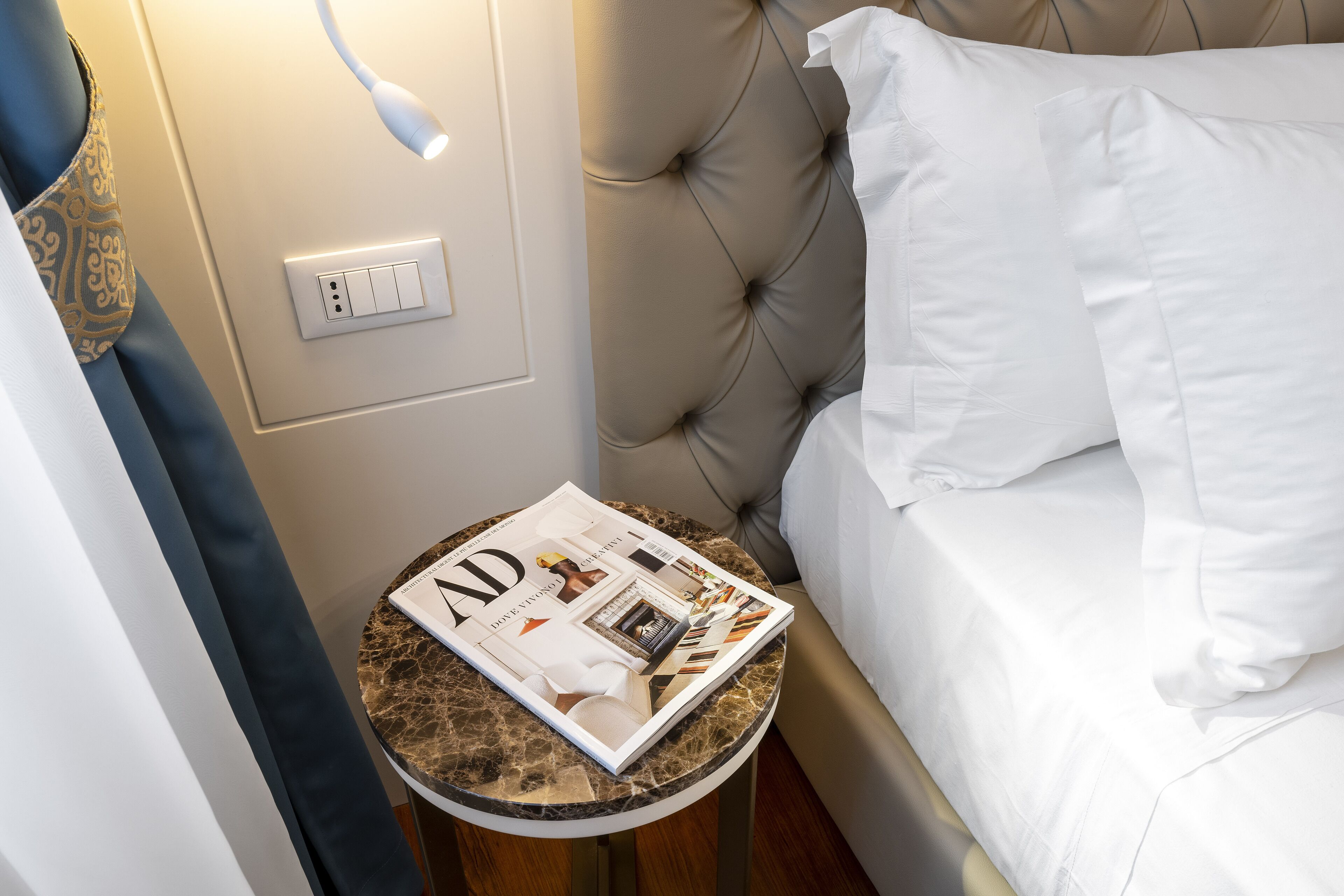A neatly made bed with white linens, a magazine on a small round table beside it, and a wall socket.
