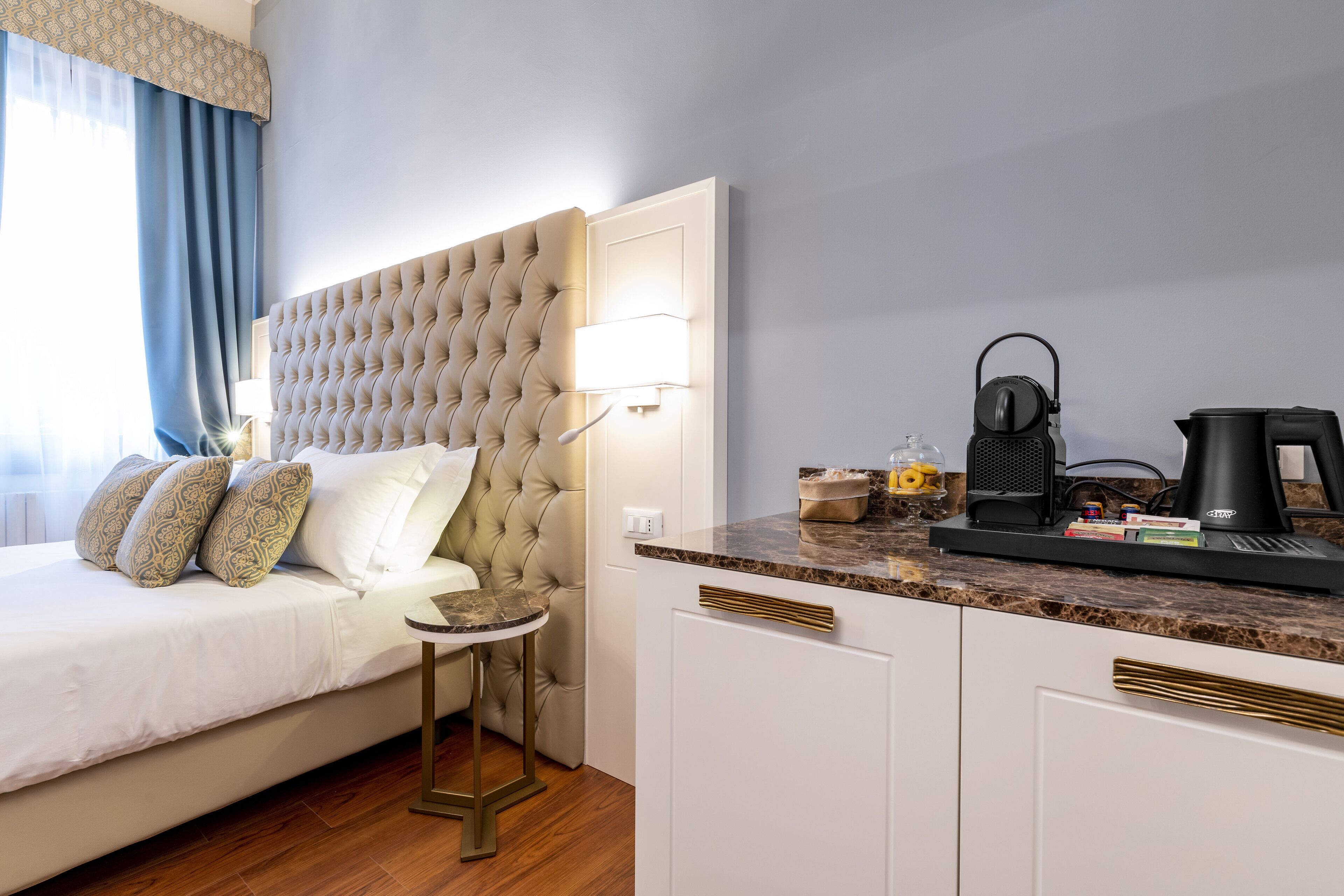 Habitació d'hotel amb un capçal de llit amb textura, una taula rodona petita, un llit amb almoinos estampats, una cafetera i una tetera a la barra.