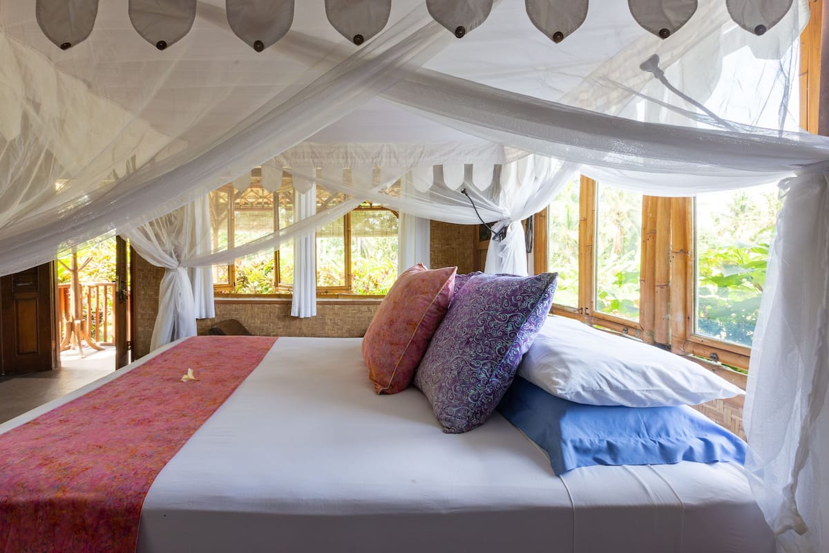 A bedroom with a large bed, white canopy, and a view of greenery outside.