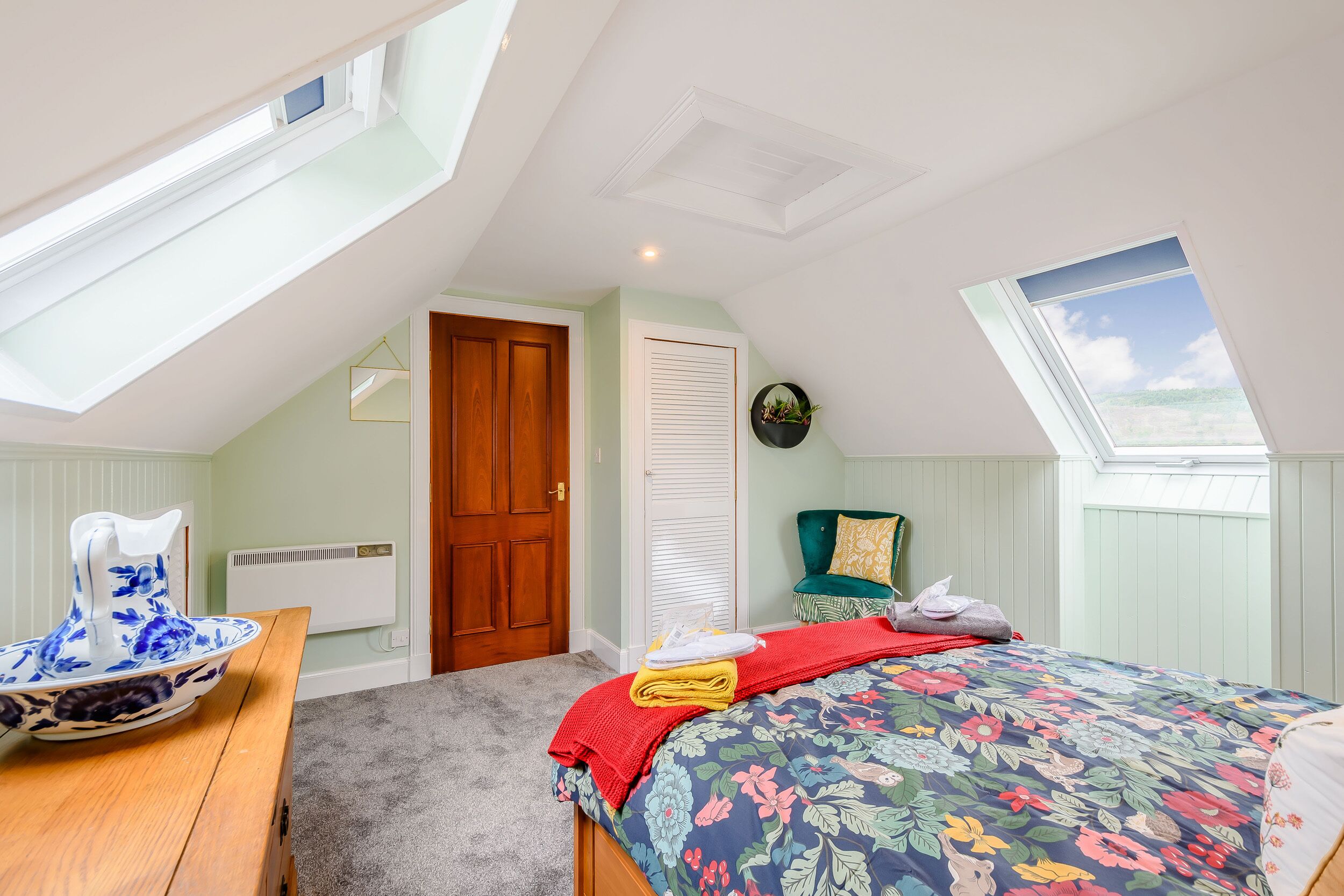 A bedroom with a skylight, a bed with a floral bedspread, a wooden table with a bowl, and a green chair.