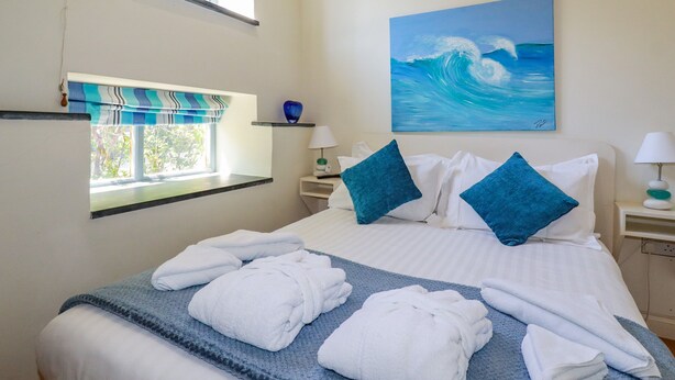 A neatly made bed with white linens and blue pillows, a painting of a wave on the wall, a window with a view of trees, and a small shelf with a blue vase.