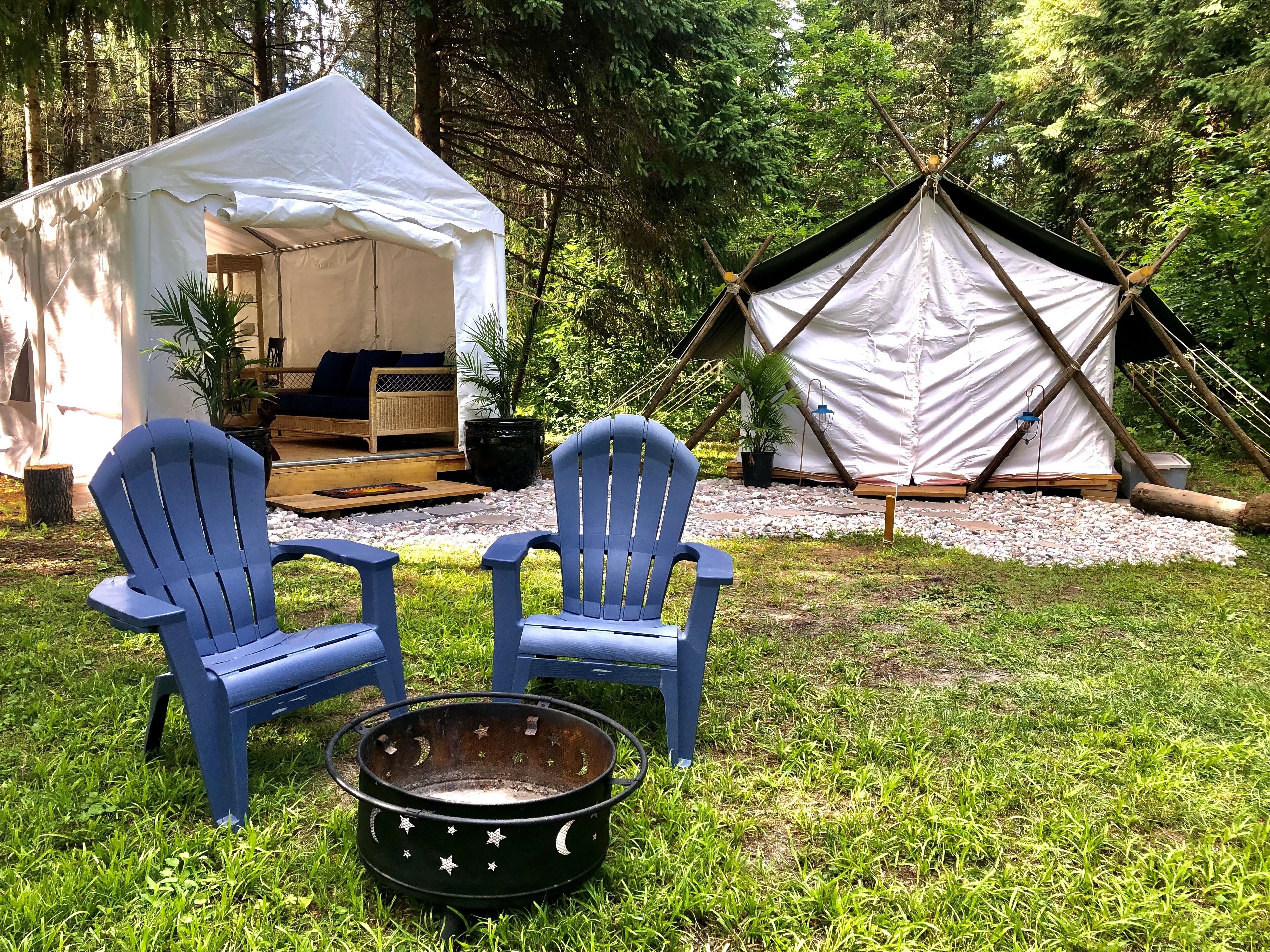 Un campamento con dos sillas Adirondack azules, un brasero negro con diseños de estrella y luna, y dos tiendas de campaña, una blanca y otra negra, ubicado en un área boscosa.
