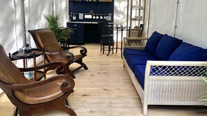 A tented room with a wooden floor, a sofa, two chairs, a small table, and a kitchen area with shelves and a hanging rack. - Glamping in the Captain’s Quarters! (Wendover)