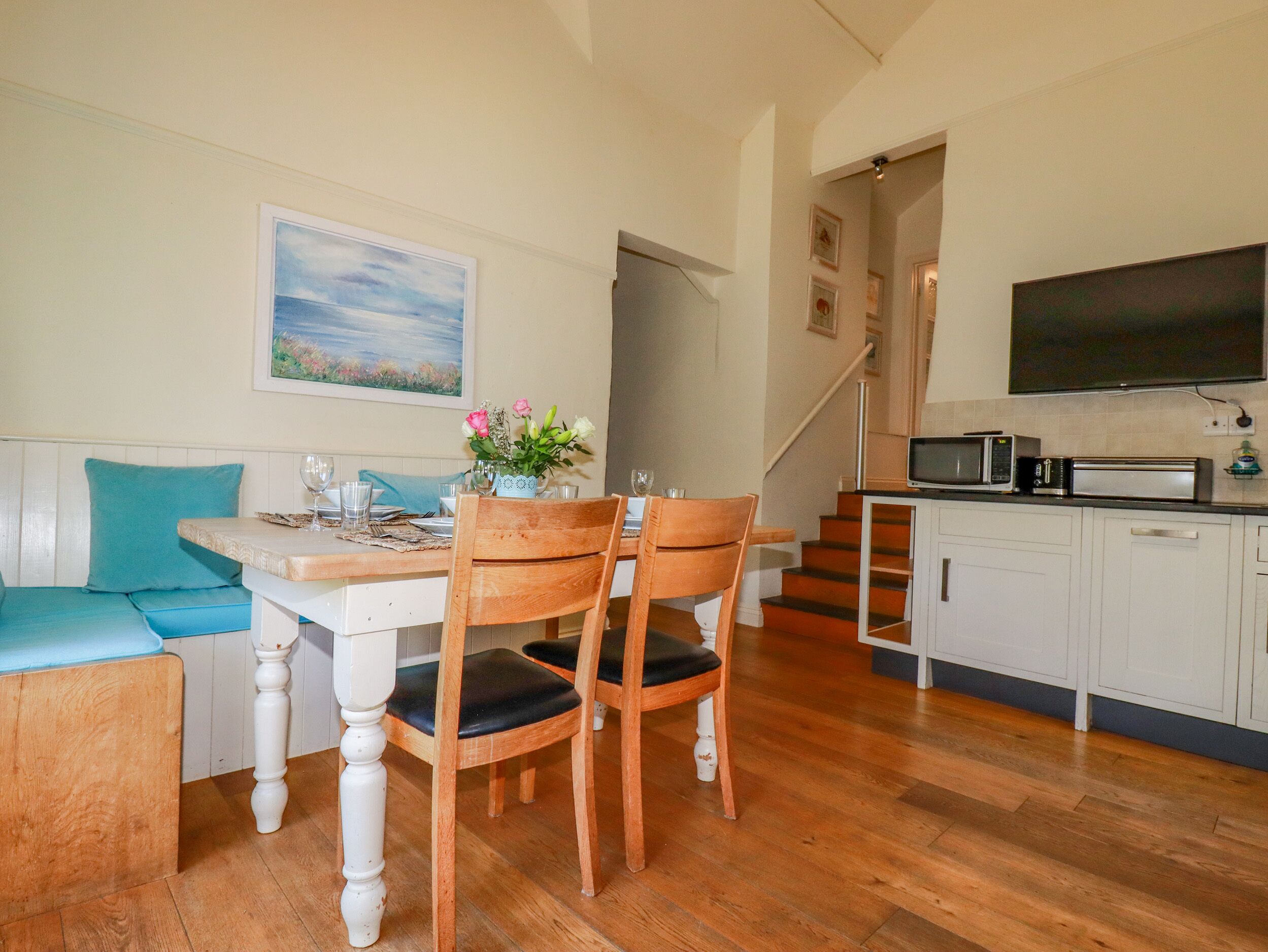 A dining area with a wooden table, chairs, and a bench. There is a kitchen with a microwave and a TV on the wall.