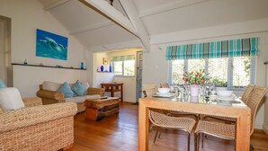 A spacious living room with a high ceiling, a dining area with wicker furniture, and a view of the outdoors through a window with a striped valance. - Abalone (Bude)