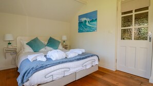 A neatly made bed with white linens and blue accents, a painting of a wave on the wall, and a wooden floor. - Abalone (Bude)