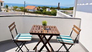 Un balcon avec une table en bois et deux chaises, un pot de fleurs, et une vue sur la mer et les maisons au loin.
