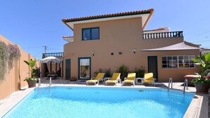 Une piscine avec des chaises longues, un patio couvert et un bâtiment à deux étages surmonté d’un toit en tuiles.