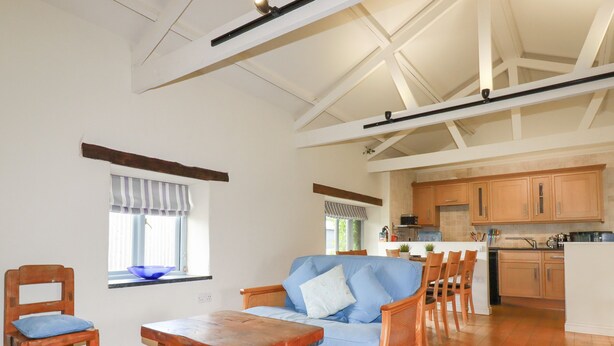 A spacious living area with a blue sofa, wooden chairs, a wooden table, and a kitchen in the background. - Oyster (Bude)