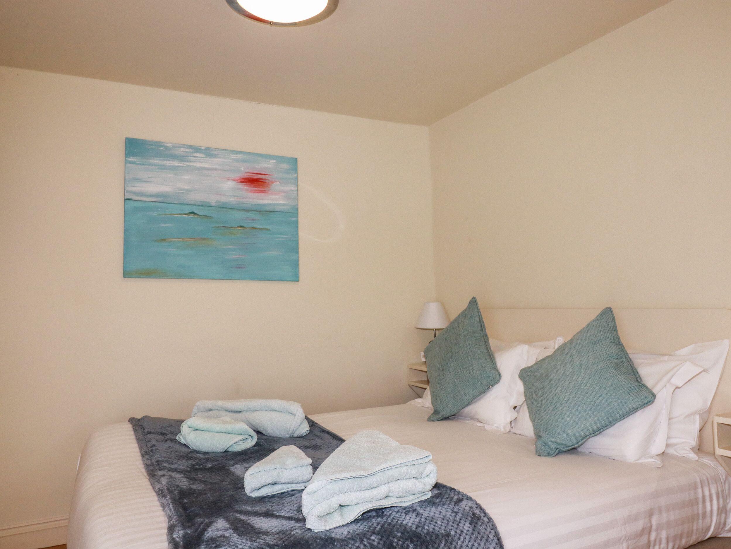 A hotel room with a bed, a painting of a seascape, and a lamp.