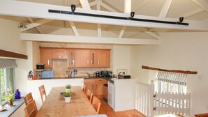 A spacious kitchen with wooden cabinets, a dining area with a wooden table and chairs, and a view of the outdoors through a window with a white fence. - Oyster (Bude)