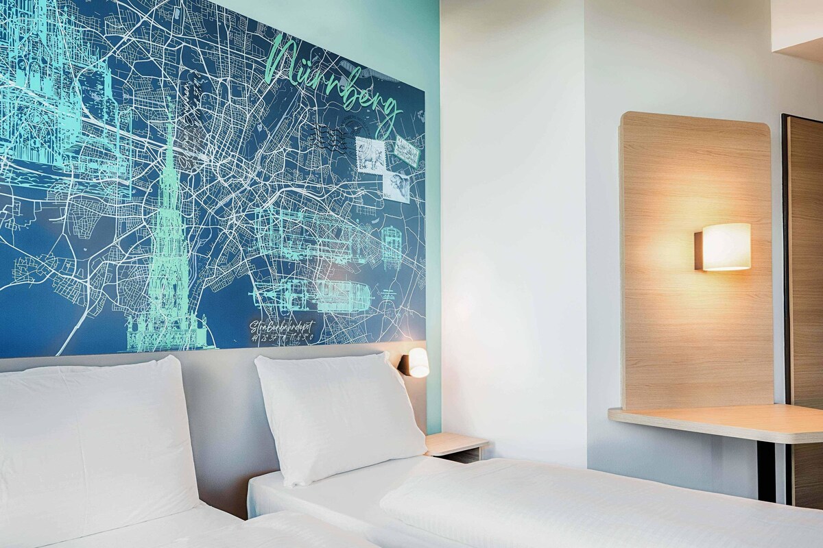 Habitación de hotel con un gran mapa de la ciudad en la pared, una cama con sábanas blancas y una mesita de noche con una lámpara.