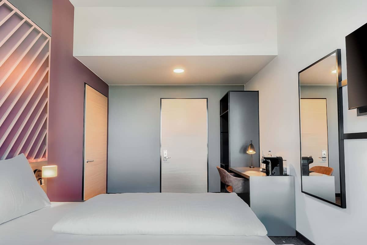 A modern hotel room with a large bed, a desk with a lamp, a chair, and a mirror.