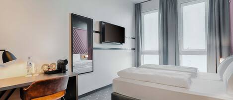 Habitación de hotel con cama, escritorio con silla, televisor montado en la pared y ventana con cortinas.