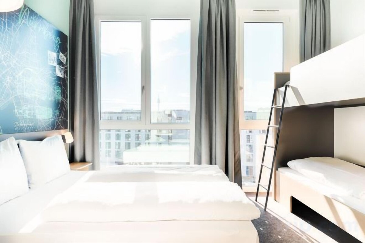 A hotel room with a large bed, a bunk bed, a window with curtains, and a view of the cityscape.