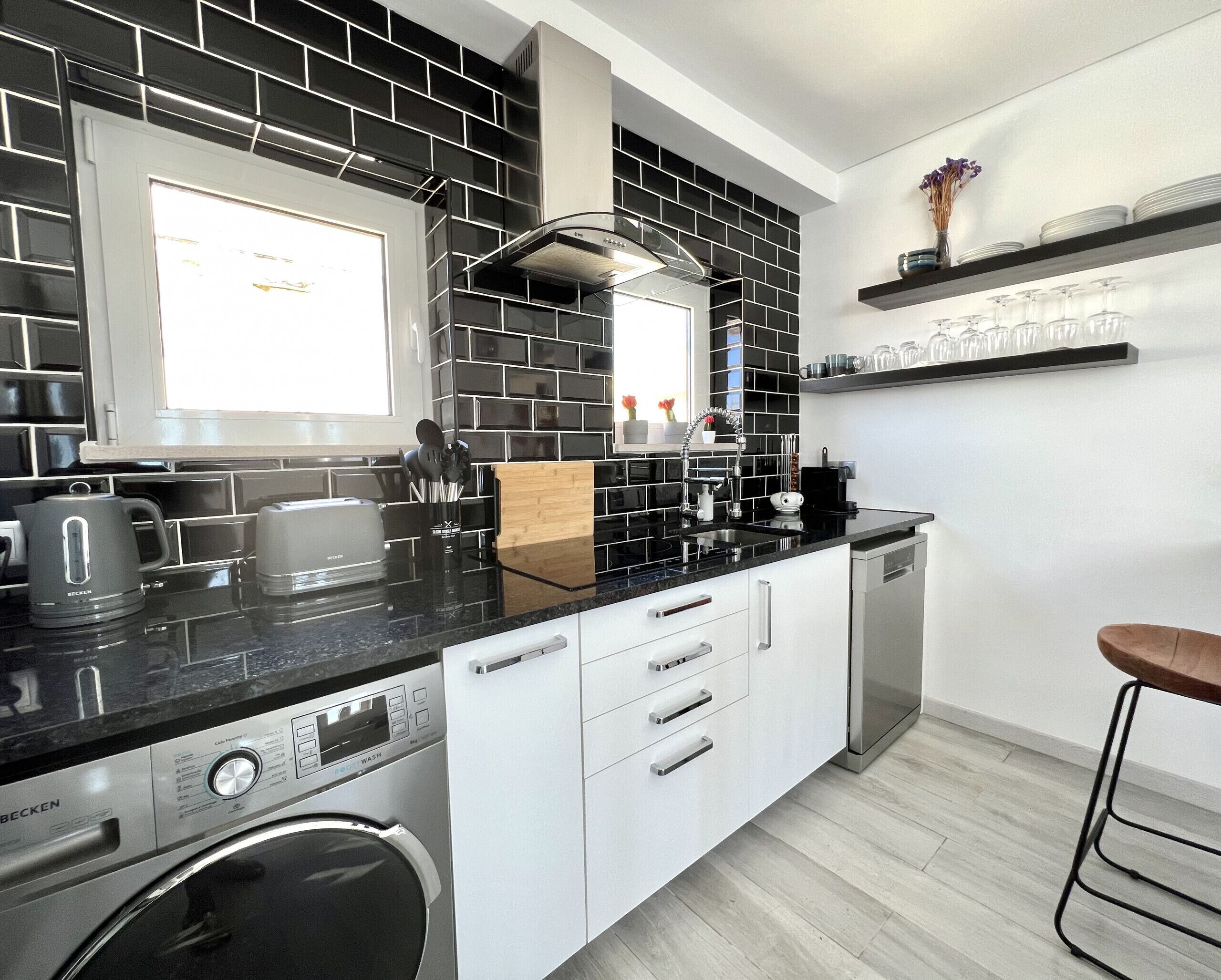 A modern kitchen with black and white tiles, a built-in dishwasher, a coffee machine, and a wooden cutting board.