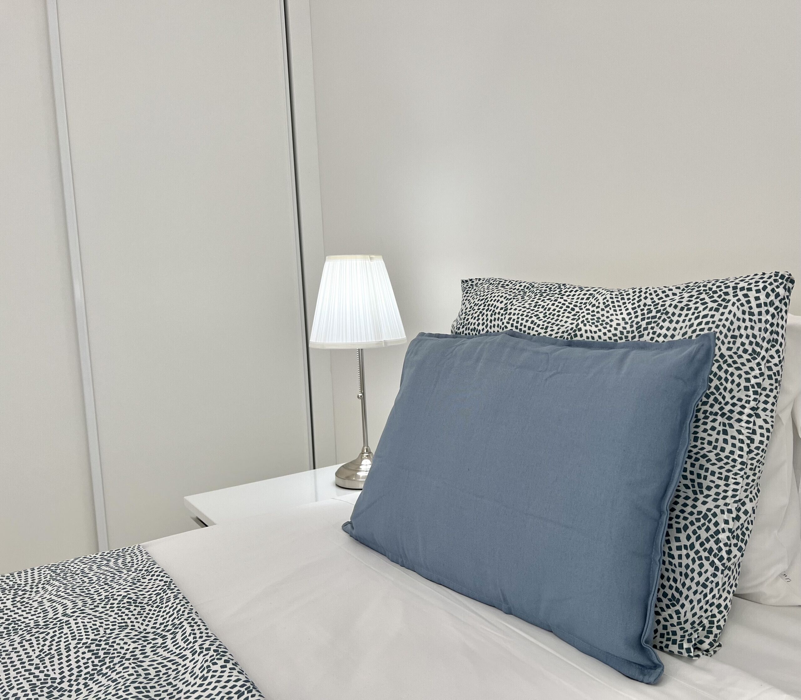 Ein ordentlich bezogenes Bett mit blauem Kopfkissen und gemusterten Zierkissen, einer Nachttischlampe und einem weißen Nachttisch.