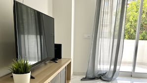 Een moderne woonkamer met een flatscreen televisie op een houten dressoir, een kleine potplant en een groot raam met dunne gordijnen.