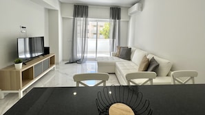 Een moderne woonkamer met een flatscreen televisie, een bank, een eettafel en een groot raam met gordijnen.