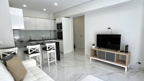 Een moderne woonkamer met een witte bank, een tv op een houten dressoir, en een keuken op de achtergrond.