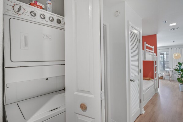 Una lavandería con lavadoras y secadoras apiladas, una puerta blanca y una cama litera en la habitación contigua.