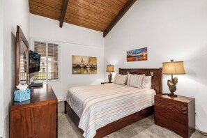 A bedroom with a wooden ceiling, a bed with a striped bedspread, two bedside tables with lamps, a dresser, a mirror, and two paintings on the wall.