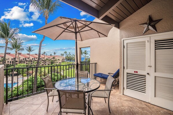 A balcony with a glass table and chairs, a beige umbrella, and a view of a pool and palm trees.