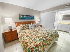 A bedroom with a bed, nightstands, a painting, and a view of a kitchen.