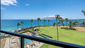 A view of a pool area with lounge chairs and a pool, a grassy lawn with palm trees, and a beach with the ocean and a mountain in the background.
